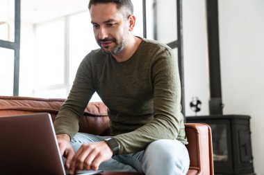Modern bir ofiste çalışan yakışıklı genç işadamı, dizüstü bilgisayar kullanıyor, kanepede oturuyor.