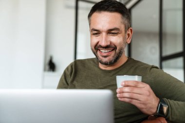 Modern bir ofiste çalışan, dizüstü bilgisayar kullanan, kanepede oturan, kahve içen yakışıklı, kendine güvenen genç bir iş adamı.