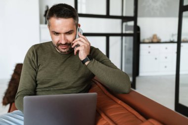 Modern bir ofiste çalışan, dizüstü bilgisayar kullanan, koltukta oturan, cep telefonuyla konuşan yakışıklı, kendine güvenen genç bir iş adamı.