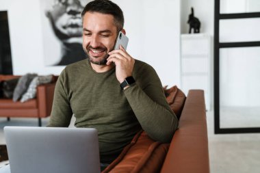 Modern bir ofiste çalışan, dizüstü bilgisayar kullanan, koltukta oturan, cep telefonuyla konuşan yakışıklı, kendine güvenen genç bir iş adamı.