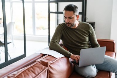 Modern bir ofiste çalışan, dizüstü bilgisayar kullanan, kanepede oturan yakışıklı, kendine güvenen genç bir iş adamı.