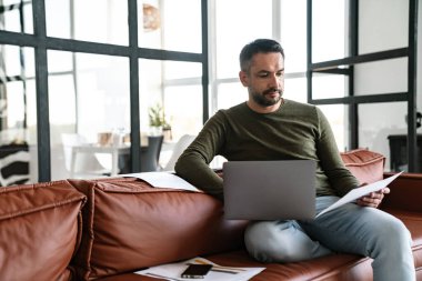 Modern bir ofiste çalışan, dizüstü bilgisayar kullanan, kanepede oturan yakışıklı, kendine güvenen genç bir iş adamı.