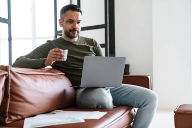 Modern bir ofiste çalışan yakışıklı, kendine güvenen genç bir iş adamı dizüstü bilgisayar kullanıyor, bir koltukta oturuyor, elinde bir fincan tutuyor.