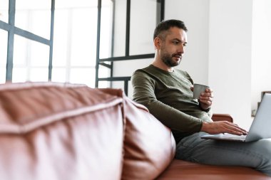 Modern bir ofiste çalışan yakışıklı, kendine güvenen genç bir iş adamı dizüstü bilgisayar kullanıyor, bir koltukta oturuyor, elinde bir fincan tutuyor.