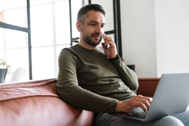 Modern bir ofiste çalışan, dizüstü bilgisayar kullanan, koltukta oturan, cep telefonu kullanan yakışıklı, kendine güvenen genç bir iş adamı.