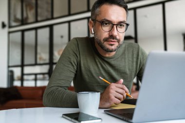 Modern bir ofiste çalışan, dizüstü bilgisayar kullanan, masada oturan, notlar alan yakışıklı, kendine güvenen genç bir iş adamı.