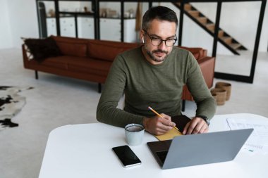 Modern bir ofiste çalışan, dizüstü bilgisayar kullanan, masada oturan, notlar alan yakışıklı, kendine güvenen genç bir iş adamı.