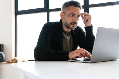 Modern bir ofiste çalışan, dizüstü bilgisayar kullanan, masada oturan yakışıklı, kendine güvenen genç bir iş adamı.