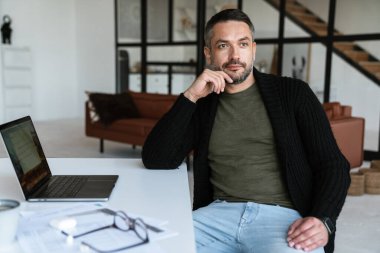 Modern bir ofiste çalışan, dizüstü bilgisayar kullanan, masada oturan yakışıklı, kendine güvenen genç bir iş adamı.