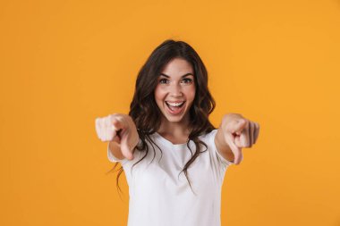 Neşeli güzel kız gülümsüyor ve sarı arka planda izole edilmiş kamerayı işaret ediyor.