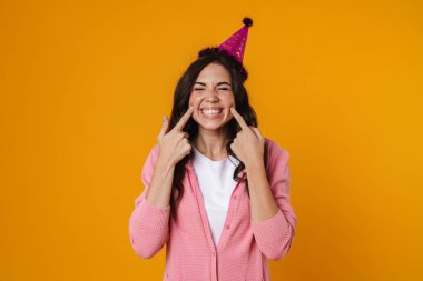 Mutlu güzel kız gülümsüyor ve sarı arka planda izole edilmiş yanaklarını işaret ediyor.