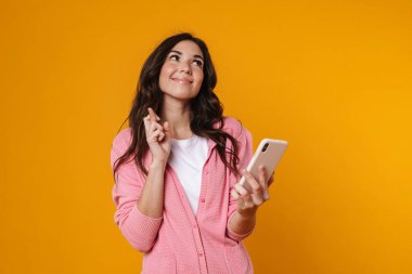 Mutlu güzel kız parmaklarını çapraz yapıp sarı arka planda izole bir cep telefonu kullanıyor.