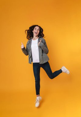 Heyecanlı güzel kız sarı arka planda tek başına zıplarken başparmağını kaldırıyor.