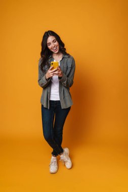 Gülümseyen ve cep telefonu kullanan neşeli güzel kız sarı arka planda izole edilmiş.