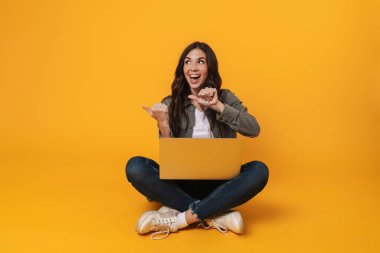Heyecanlı kız sarı arka planda izole bir şekilde otururken dizüstü bilgisayar kullanıyor.