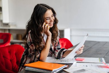 Güzel bir kadın cep telefonuyla konuşuyor ve kafede kahve içerken gazetelerle çalışıyor.