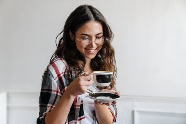 Gözlüklü, mutlu, esmer kadın içeride kahve içiyor ve gülümsüyor.