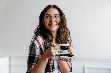 Gözlüklü, mutlu, esmer kadın içeride kahve içiyor ve gülümsüyor.