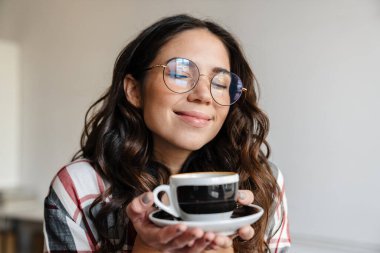 Gözlüklü, mutlu, esmer kadın içeride kahve içiyor ve gülümsüyor.