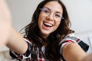 Gözlüklü neşeli bir kadın gülümsüyor ve evde selfie çekiyor.
