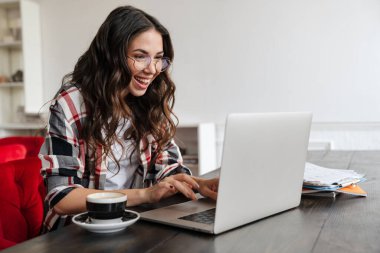Gülümseyen ve kafede kahve içerken dizüstü bilgisayarla çalışan mutlu bir kadın.