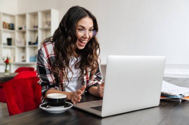 Gülümseyen, laptopla çalışan ve kafede kahve içen heyecanlı bir kadın.