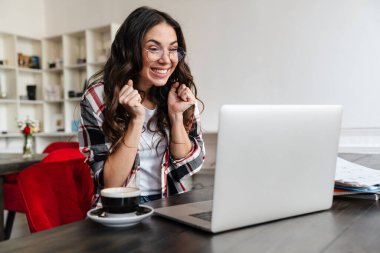 Gülümseyen, laptopla çalışan ve kafede kahve içen heyecanlı bir kadın.