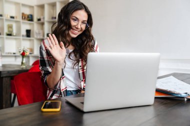 Neşeli güzel bir kadın el sallıyor ve kafede otururken dizüstü bilgisayar kullanıyor.