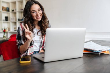 Neşeli hoş bir kadın barış işareti yapıyor ve kafede otururken dizüstü bilgisayar kullanıyor.