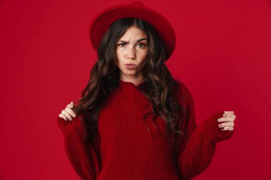 Mutsuz, güzel, şapkalı, poz veren ve kırmızı arka planda izole edilmiş kameraya bakan esmer kız.