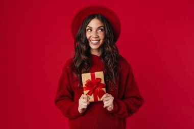 Gülen ve poz veren şapkalı neşeli, güzel esmer kız kırmızı arka planda izole edilmiş hediye kutusuyla