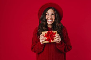 Gülen ve poz veren şapkalı neşeli, güzel esmer kız kırmızı arka planda izole edilmiş hediye kutusuyla