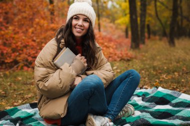 Bir sonbahar parkında elinde kitapla oturan mutlu ve çekici genç bir kadın.
