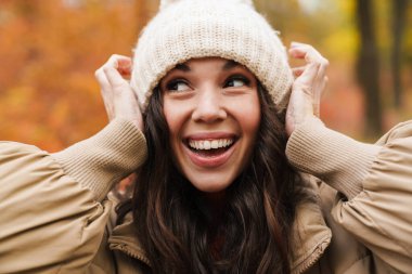 Güzel, neşeli bir kadın gülerek ve örgü şapkayla dalga geçerek sonbahar ormanlarında gezinirken