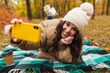 Esmer ve mutlu kadın sonbahar ormanlarında dinlenirken cep telefonuyla selfie çekiyor.