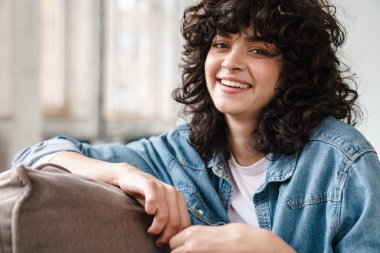 Evdeki kanepede otururken kameraya bakıp gülümseyen güzel kıvırcık kız.