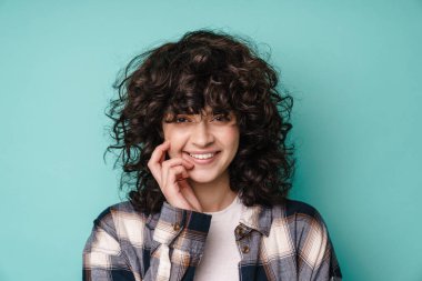 Neşeli, güzel kıvırcık kız gülümseyen ve mavi arka planda izole edilmiş kameraya bakan.