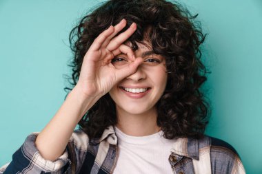Neşeli, güzel, kıvırcık kız gülümseyen ve işaret eden tamam işareti mavi arka planda izole edilmiş.