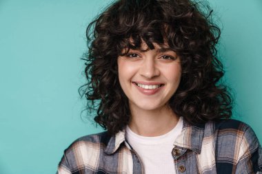 Neşeli, güzel kıvırcık kız gülümseyen ve mavi arka planda izole edilmiş kameraya bakan.