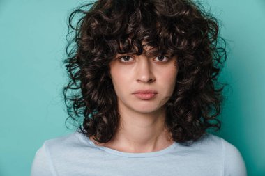 Beyaz, ciddi kıvırcık kız. Mavi arka planda izole edilmiş kameraya bakıyor ve poz veriyor.