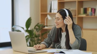 Kulaklık takan gülümseyen Asyalı genç bir Koreli ofiste dizüstü bilgisayarını kullanıyor.