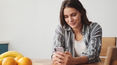 Güzel bir genç kadın akıllı telefonunu evde masada otururken kullanıyor.