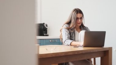 Gözlük takan odaklanmış genç bir kadın evdeki mutfaktaki masada dizüstü bilgisayarıyla çalışıyor.