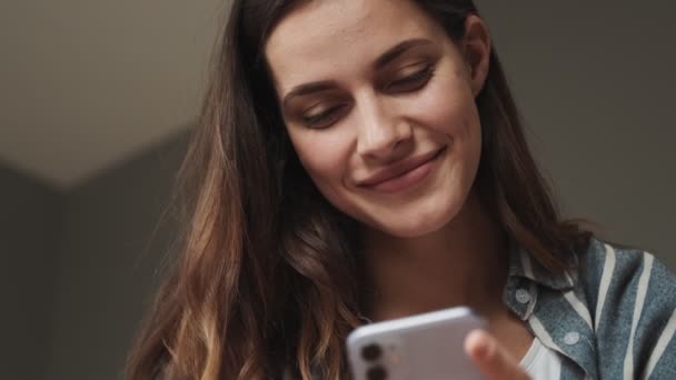 笑顔の若い女性のクローズアップビューは 自宅の寝室に座って彼女のスマートフォンを使用しています — ストック動画