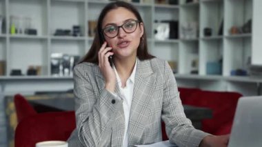 Gözlüklü genç bir iş kadını, dairelerde dizüstü bilgisayarıyla çalışırken cep telefonuyla konuşuyor.