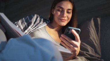 Memnun genç bir kadın akıllı telefonunu kullanıyor. Dairenin içinde oturuyor.