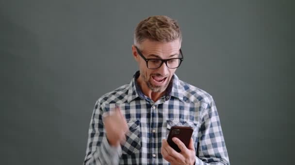 一个戴着眼镜的快乐的成年人在工作室里用他的智能手机在灰色背景下孤立无援地做着获胜的手势 — 图库视频影像