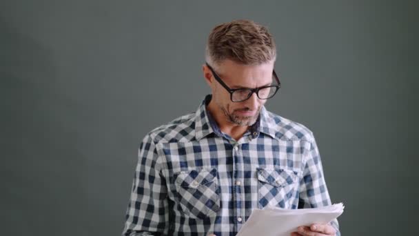 一个戴眼镜的专注的成年人拿着纸质文件 站在工作室灰色的背景下孤立地看着它们 — 图库视频影像