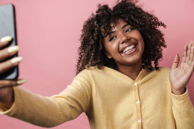 Komik Afro-Amerikalı kadın pembe duvarın üzerinden izole edilmiş cep telefonuyla selfie çekerken el sallıyor.