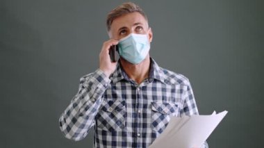 Koruyucu maske takan iyi görünümlü yetişkin bir adam, stüdyoda gri bir arkaplanda tek başına duran akıllı telefonla konuşurken kağıt belgelere bakıyor.